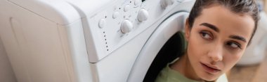 Young woman looking at washing machine in laundry room, banner 