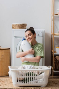 Positive woman holding washing liquid and looking at camera in laundry room 