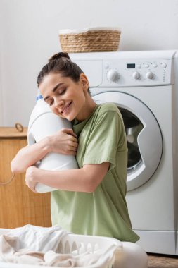 Cheerful young woman hugging bottle with washing liquid near basket with clothes in laundry room 