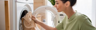 Side view of brunette woman putting clothes in washing machine, banner 