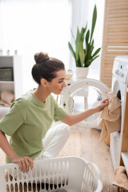 Side view of young woman putting clothes in washing machine near basket in laundry room 