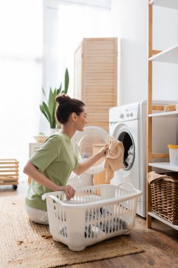 Side view of smiling woman putting clothes in washing machine at home 