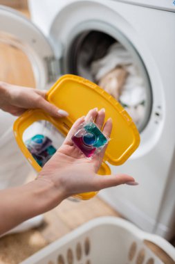 Cropped view of woman holding detergent pod and box near machine in laundry room 