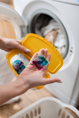 Cropped view of woman holding detergent pod and box near machine in laundry room  clipart