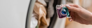 Cropped view of woman holding detergent pod near blurred washing machine in laundry room, banner 