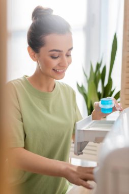 Smiling woman holding washing liquid near machine in laundry room 