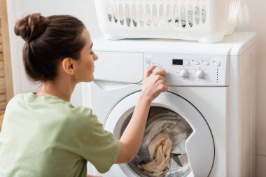 Side view of blurred woman switching washing machine near basket in laundry room  clipart