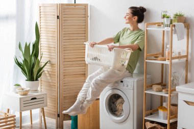 Side view of young woman holding basket while sitting on washing machine in laundry room 