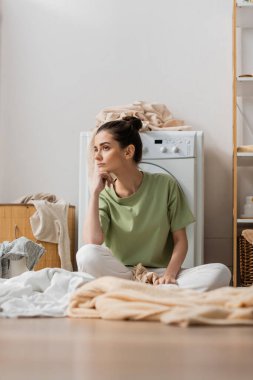 Sad woman sitting near clothes on floor in laundry room 