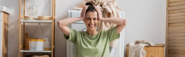 tensed woman touching head near washing machine in laundry room, banner  clipart