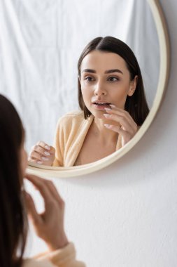 Brunette woman in bathrobe looking at mirror in bathroom in morning 