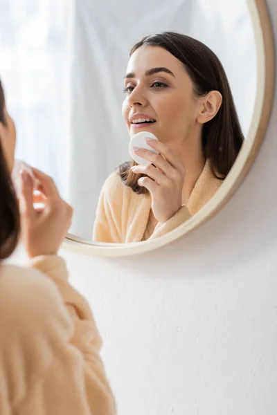 reflection of brunette young woman cleansing face with cotton pad in bathroom mirror