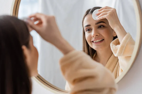 reflection of smiling young woman cleansing face with cotton pad in bathroom mirror