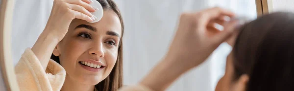 reflection of smiling young woman cleansing face with cotton pad in bathroom mirror, banner