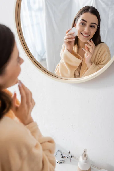 reflection of pleased young woman cleansing face with cotton pad in bathroom mirror