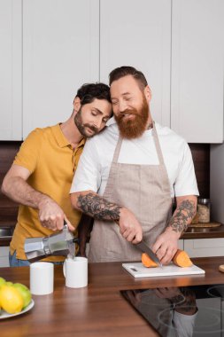 Gay man pouring coffee near smiling husband cutting sweet potato in kitchen 