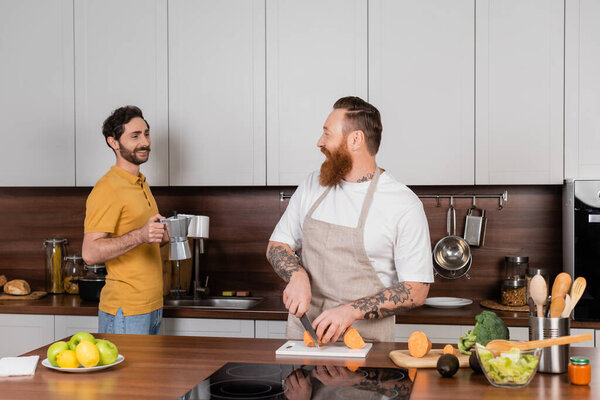 Smiling gay man holding coffee and cups near husband cooking in kitchen 