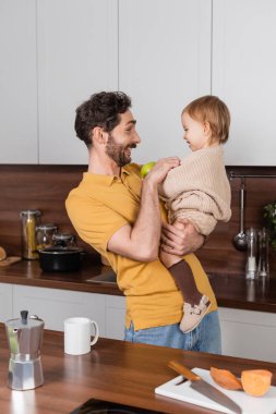 Gülümseyen bir adamın, yeni yürümeye başlayan kızını mutfakta elmayla tutuşu. 