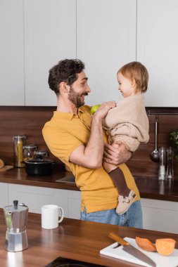 Side view of smiling man holding toddler daughter with apple in kitchen 