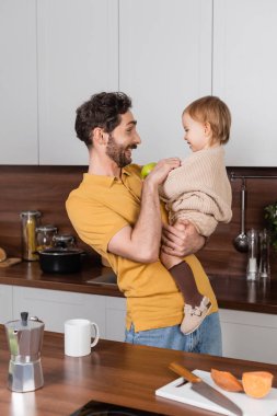 Side view of smiling man holding toddler daughter with apple in kitchen  clipart