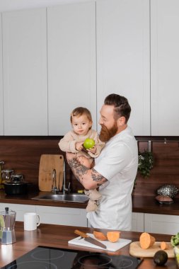 Bearded man holding toddler daughter with apple in kitchen at home 