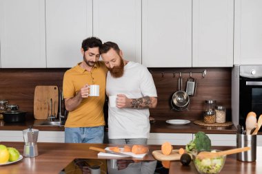 Gay couple holding cups of coffee near vegetables in kitchen 