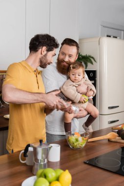 Gülümseyen gay, yeni yürümeye başlayan kızını kucağına alıp mutfakta salata pişiriyor. 