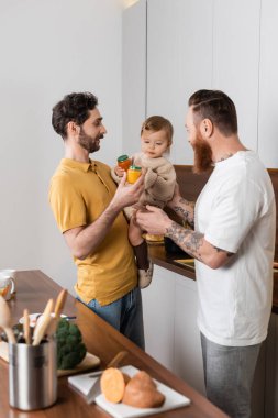 Homosexual couple holding baby food near toddler girl in kitchen 