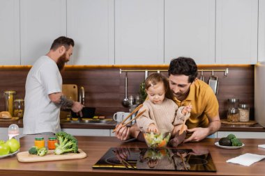 Eşcinsel adam mutfakta taze salatanın yanında elinde kaşıkla küçük kızını tutuyor. 
