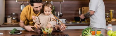 Homosexual parent holding daughter with spoon and fresh salad in kitchen, banner  clipart