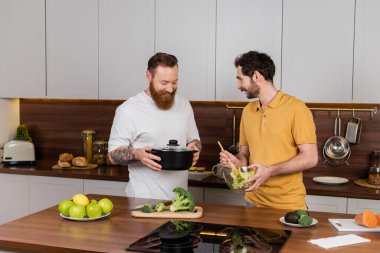 Tattooed gay man holding pot near partner with fresh salad in kitchen 