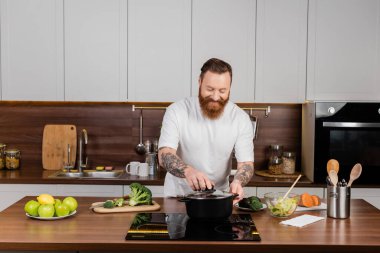 Bearded man putting cap on pot while cooking in kitchen  clipart