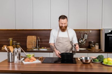 Tattooed man looking at pot on stove while cooking in kitchen 