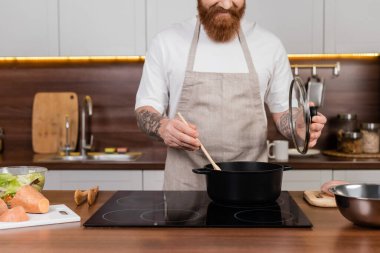 Mutfaktaki salatanın yanında sakallı bir adamın tencerede yemek pişirmesi. 