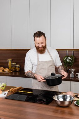 Smiling bearded man holding pot near stove and food in kitchen 