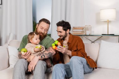 Homosexual couple holding toy near baby daughter with fresh apple on couch at home clipart