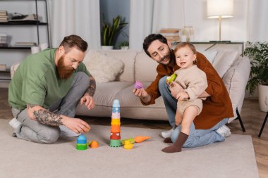 Smiling gay couple playing with baby daughter holding apple at home 