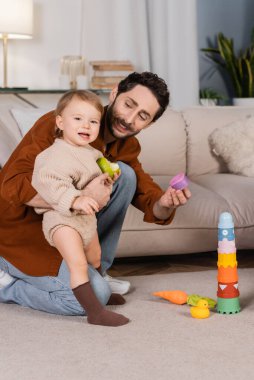 Smiling dad holding toy near baby daughter with apple at home 