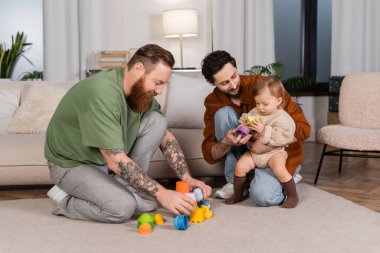 Same sex parents holding toys near baby girl with apple at home 