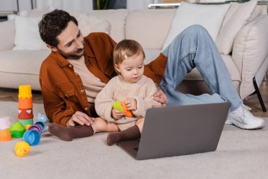 Father sitting near baby daughter with toy and laptop at home 