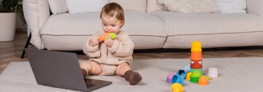 Toddler girl holding toy and looking at laptop at home, banner 