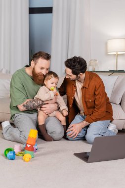 Gay couple hugging daughter near toys and laptop at home  clipart
