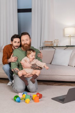 Same sex couple with toddler daughter looking at camera near toys and laptop in living room  clipart