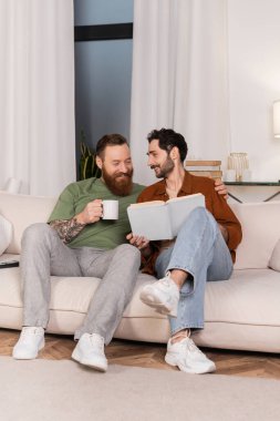 Cheerful same sex couple holding coffee and book on couch at home 