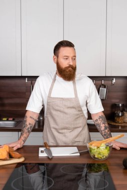 Önlüklü dövmeli adam mutfakta salatanın yanında duruyor. 