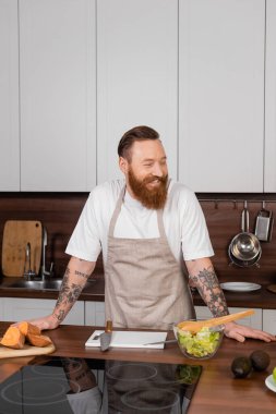 Cheerful tattooed man in apron standing near food and fresh salad in kitchen 
