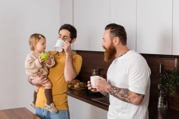 Gay couple drinking coffee near toddler daughter with apple in kitchen