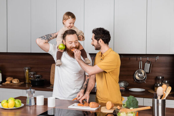 Gay man feeding partner near baby daughter in kitchen 