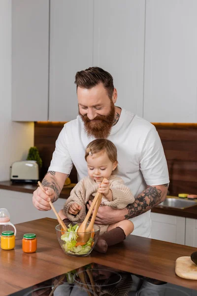 Bearded dad cooking fresh salad near toddler daughter in kitchen