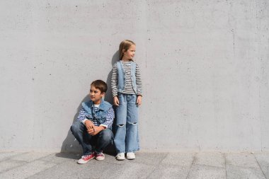 full length of stylish kids in denim outfits with striped long sleeve shirts posing near grey wall in mall  clipart